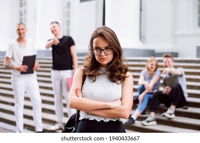 Angry And Upset College Girl Standing With Her Hands Crossed, Being Bullied By Her Group Mates. College Students Mocking On New Learner.