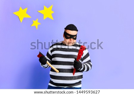 angry and upset fat young offender looking pensively at pipe wrench, making decision, choosing a tool for breaking the house abuse, violence and bullying concept. isolated blue background. studio shot