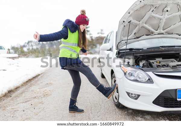 Angry woman kicking her broken car at roadside.\
Winter scene.