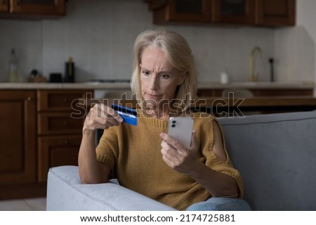 Angry worried older woman sits on sofa at home holds debit card and cellphone experience lack of money to make e-payment due to insufficient funds, suspicious unauthorized withdrawal from card concept