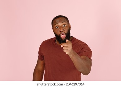 Angry young Afro-American guy with rolled eyes in brown t-shirt points finger of blame to camera on studio pink background closeup