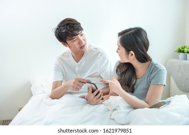 Angry Young Asian Couple Or Marriage Fighting For A Mobile Phone At Home. Jealous Caucasian Woman Holding Smart Phone And Showing Message To His Husband With Argument And Unsatisfied Motion.