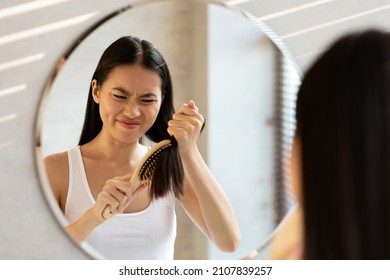 Angry young asian woman having problem of her thick hair while using hair brush, looking at mirror and combing hair, bathroom interior, mirror reflection shot. Dry, frizzy, damaged hair concept