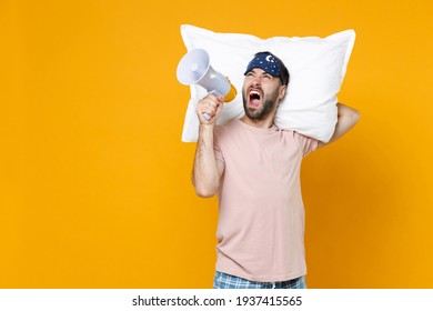Angry young bearded man 20s in pajamas home wear sleep mask hold pillow screaming in megaphone while resting at home isolated on yellow background studio portrait. Relax good mood lifestyle concept