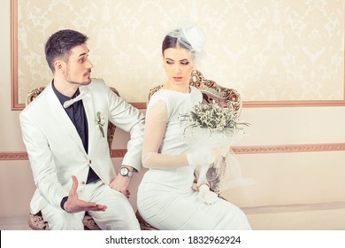 Angry young bride and groom, man and woman upset on each others sitting in armchairs looking to the side isolated on classic background. Conflict between bride and groom concept