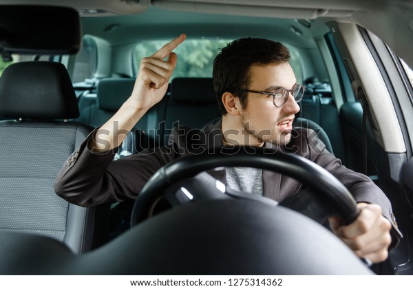 Angry young driver quarrels with other\
drivers. His left hand is on the steering\
wheel.