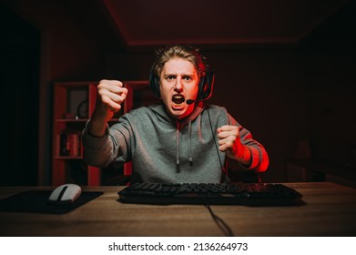 Angry young male gamer in headset looks at screen and shows fist during intense game. Portrait of an emotional gamer.
