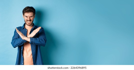 Angry young man frowning and clenching teeth outraged, showing cross gesture to stop or forbid something, standing on blue background. - Shutterstock ID 2264396895