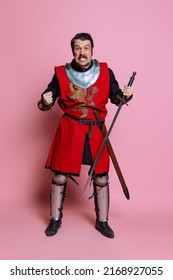 Angry Young Man, Medieval Warrior Or Knight Wearing Wearing Armor Clothing Posing Isolated Over Pink Background. Model With Wounded Face. Comparison Of Eras, History, Funny Meme Emotions