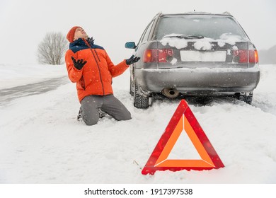 Angry young man in an orange jacket is kneeling on a snow-covered road. He is in a state of stress because his car is broken. Winter travel and recreation, road problems and assistance concepts