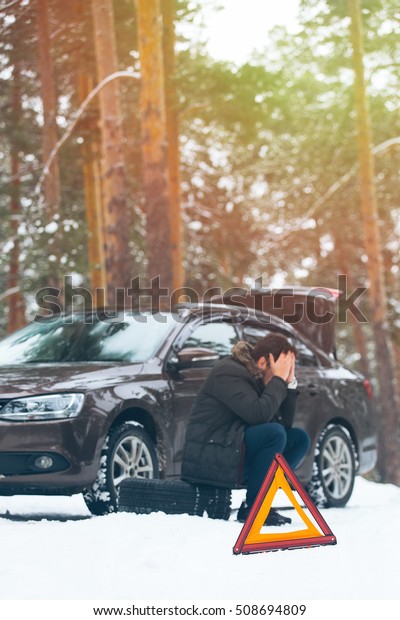 Angry young man\
waiting for help, sitting near the broken car on the side of the\
road in the winter in the\
woods