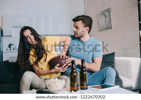 angry young woman fighting for american football ball with husband during match on tv at home