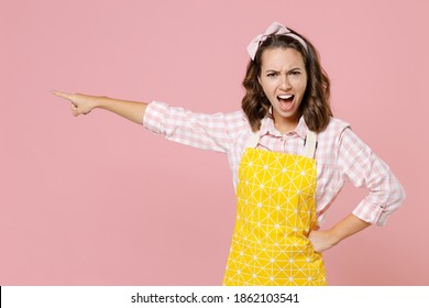 Angry young woman housewife 20s in yellow apron pointing index finger aside screaming swearing while doing housework isolated on pastel pink colour background studio portrait. Housekeeping concept