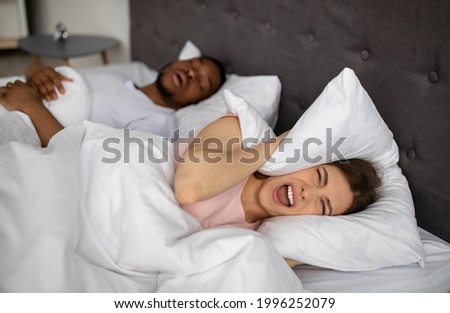 Angry young woman shouting and covering ears with pillow while her husband snoring on bed. Millennial lady being disturbed by her boyfriend's snort problem, cannot fall asleep