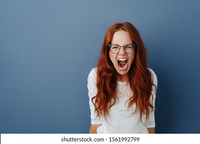 Angry Young Woman Throwing A Temper Tantrum Yelling At The Camera With A Furious Expression Over A Blue Studio Background With Copy Space