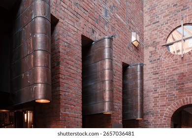 An angular view of a modern red brick building's exterior at night. The facade features large, curved copper light fixtures with a green patina, casting a warm glow downwards.