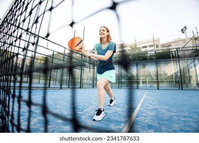 Angular view of a photo through a net of a paddle court of a cute girl in sportswear posing with a racket waiting to receive a serve. Concept of women playing paddle. Paddle for women.