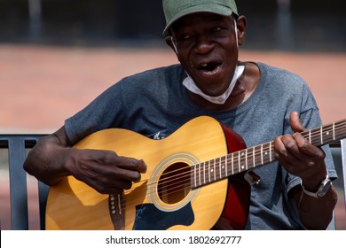 Annapolis, MD 08/21/2020: An Elderly African American Man Wearing Baseball Hat And A Face Mask Is Playing Classical Guitar And Singing While Sitting On A Bench On A Street. He Is A Street Musician.