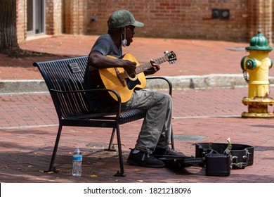 Annapolis, MD 08/21/2020: An Elderly African American Man Wearing Baseball Hat And A Face Mask Is Playing Classical Guitar And Singing While Sitting On A Bench On A Street. He Is A Street Musician.