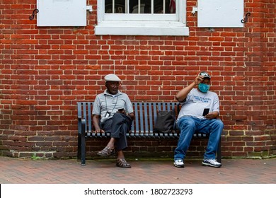 Annapolis, MD 08/21/2020: Two Elderly African American Men Are Sitting On A Bench Outside An Old Brick Building In Annapolis. One Wears Face Mask Due To COVID-19. They Both Look At Their Cell Phones.