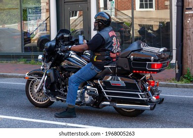 Annapolis, MD, USA 05-02-2021: An African American Man With Goatee Beard Wearing Jeans, Converse Shoes, Vest And Helmet Is Idling On The Road In The City. He Is Riding A Harley Davidson Motorbike.