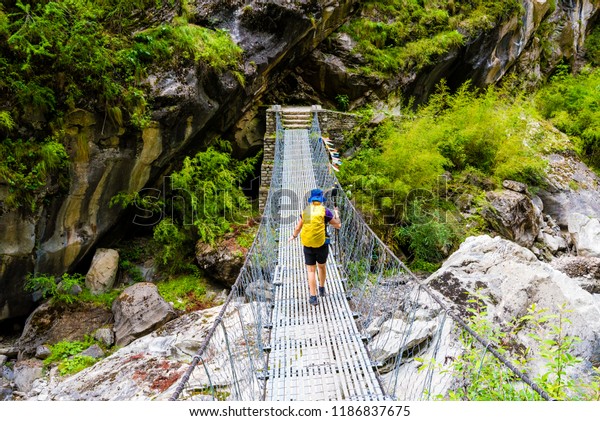 Annapurna Conservation Area, Nepal - July 19, 2018\
: Woman backpacker on trekking path crossing a suspended bridge in\
Annapurna Conservation Area, a hotspot destination for mountaineers\
in Nepal