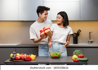 Anniversary Gift. Asian Couple Celebrating Family Holiday, Husband Giving Wrapped Present Box To Happy Wife Surprising Her Standing In Modern Kitchen At Home. Celebratory Dinner Preparation