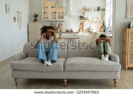 Annoyed angry African American mother sitting with offended upset son on sofa at home, talking on phone, discussing child behavior with spouse. Mom and kid conflict. Parents, children and discipline