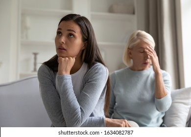 Annoyed Angry Millennial Adult Girl Sit On Couch Separate From Distressed Senior Mom Thinking Of Family Problems Or Misunderstanding, Pensive Grownup Daughter Having Argument With Middle-aged Mother