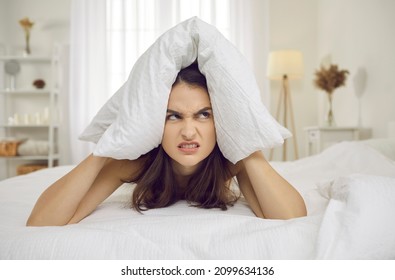 Annoyed and angry woman closes her ears with pillow in morning because of noise. Close up portrait of sleepless woman annoyed by alarm clock or loud neighbors. Concept of insomnia, stress and tinnitus