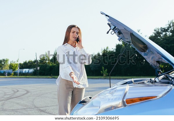 An\
annoyed and angry woman, near a broken car, tries to call an\
insurance agent and a repair crew. for car\
repair