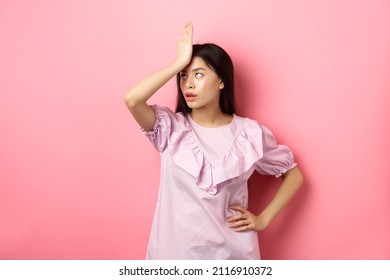 Annoyed asian girl roll eyes and facepalm, feeling tired and irritated with stupid conversation, standing against pink background