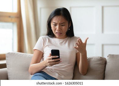 Annoyed Asian millennial girl sit on couch in living room frustrated with slow Internet connection or spam on smartphone, mad Vietnamese young woman confused with cellphone device operational problem - Powered by Shutterstock