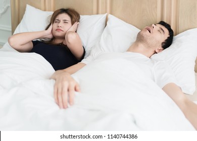 Annoyed Asian wife blocking her ears from noise of husband snoring in bedroom at home. Young couple have problem with man's snoring.