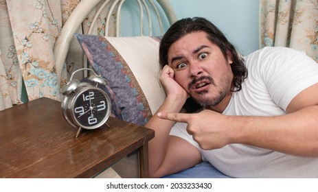 An Annoyed Bearded Man Of Mixed Race Points To An Alarm Clock At His Bedside Which Did Not Ring Or Too Noisy.