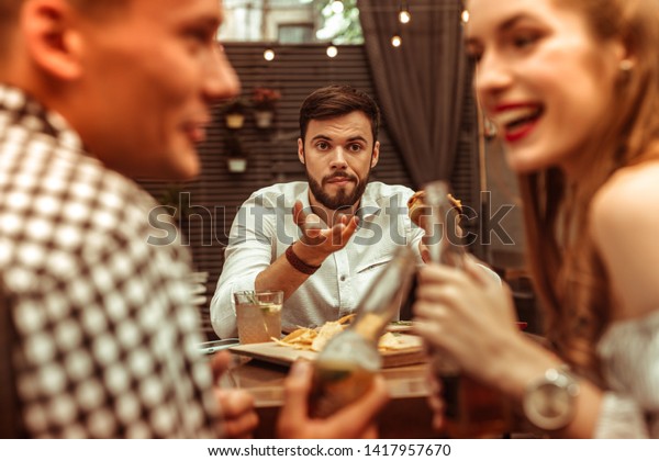 Annoyed bored man. Handsome
short-haired nice-appealing young-adult annoyed bored man holding a
burger and feeling lonely while his friends are flirting with each
other