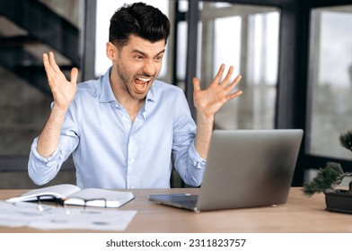 Annoyed caucasian office employee, freelancer or broker sitting at a table in a modern office, indignantly looking at a laptop, overworked, stressed, gesturing with his hands, shouting - Shutterstock ID 2311823757