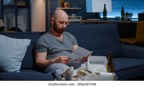 Annoyed, Desperate Man Sitting On Sofa Reading Eviction Notice In Letter Ripping Warning Document Crying Overwhelmed By Problems. Shocked, Worried Tenant By Warning Notification For Unpaid Bank Bills