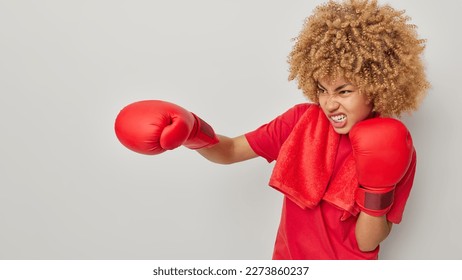 Annoyed displeased curly haired sportswoman clenches teeth makes punch wears boxing gloves dressed in red t shirt isolated over grey background copy space aside. Kickboxing concept. Dynamic movement