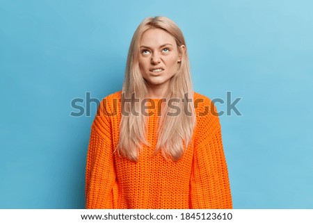 Annoyed dissatisfied woman concentrated above smirks face and looks at something with aversion has long blonde hair dressed in knitted orange jumper isolated over blue wall. Negative face expressions