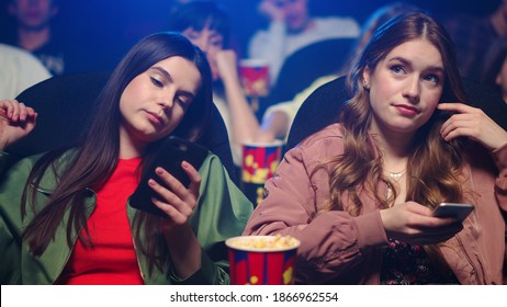 Annoyed Female Friends Looking To Cellphones In Movie Theater. Upset Girls Checking Smartphone In Cinema. Disappointed Women Scrolling Mobile Phone Indoor