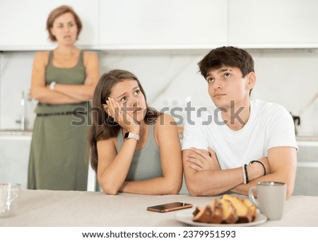 Annoyed female scolds son and daughter-in-law for infantile misconduct unworthy behavior. Young couple girl and guy are sitting at table bored and wearily of talking to irritated and screaming mother