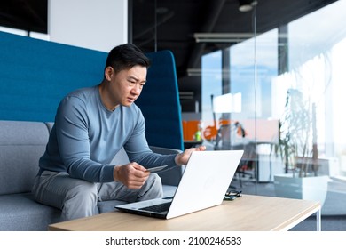 Annoyed And Frustrated Asian Man Trying To Make An Online Purchase Sitting In The Office At The Computer, Using A Credit Card