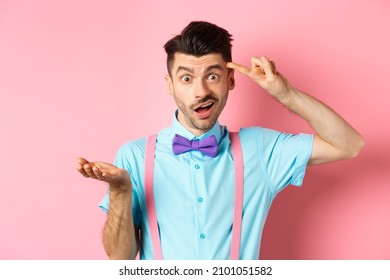 Annoyed guy mocking someone stupid, pointing finger at head and camera, are you stupid gesture, standing over pink background