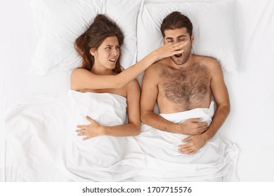 Annoyed Irritated European Wife Covers Husbands Nose, Cannot Sleep Because Of Loud Snoring, Feels Bad, Has Sleepless Night, Pose In Comfortable White Bed. Sleeping Problems And People Concept