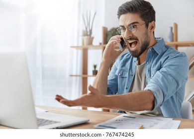 Annoyed Male Enterpreneur Looks Angrily Into Laptop Computer, Has Conversation With Business Partner Via Mobile Phone, Being Annoyed As He Didn` T Prepare Business Report For Meeting With Investors