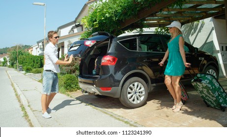 Annoyed Man Sees His Girlfriend Bringing Out More Luggage In Front Of Their House To Pack In Black Car. Young Woman Carrying Travel Bag Smiles While Walking Towards Her Husband Standing In Driveway.