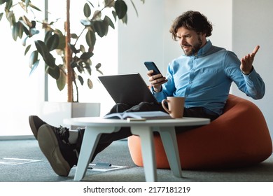 Annoyed Mature Businessman With Laptop Looking At Cellphone Screen And Gesturing Angrily, Working At Office, Copy Space. Frustrated Boss Having Problems With Project, Speaking With Employee By Phone