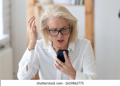 Annoyed mature businesswoman seated at workplace desk gesticulates holding smart phone looks at screen feels indignant wi-fi connection problems, business app crashes, synchronization failure concept