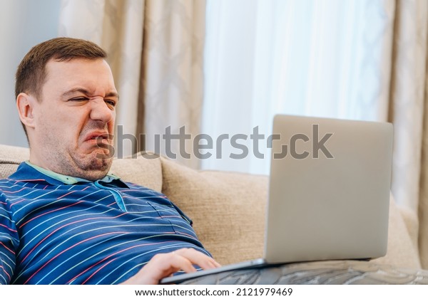 Annoyed millennial man with disgust confused looking\
at laptop screen sitting on sofa at living room. Cringe guy looking\
at computer with squeamishness, reacting on watching disgusting\
video at home